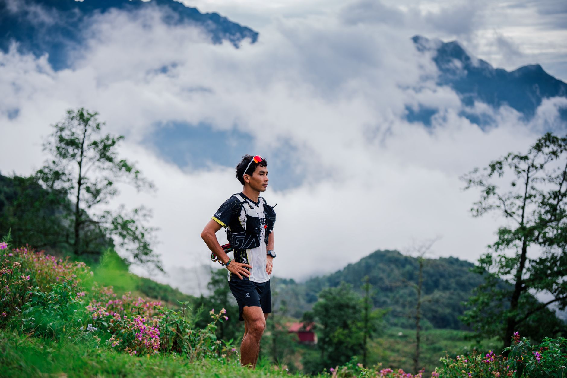 Trail runner pausing in lush green mountains with clouds and flowers.